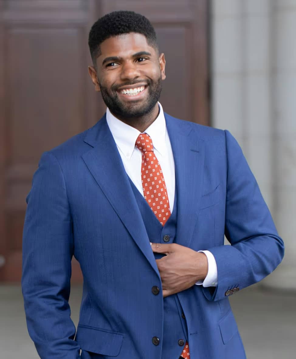 Smiling man in a blue suit with a white shirt and red patterned tie, standing with one hand adjusting his jacket.