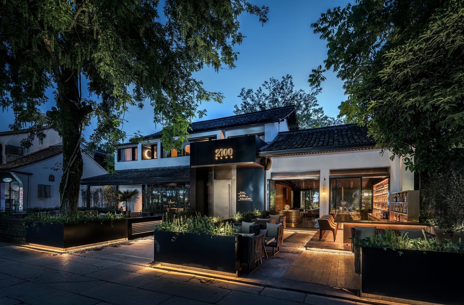 Exterior of a modern restaurant with outdoor seating and warm lighting at dusk, surrounded by trees.