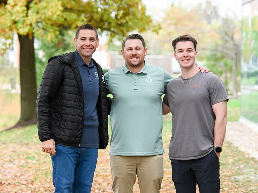 Three men standing outdoors in a park-like setting, smiling at the camera on an autumn day