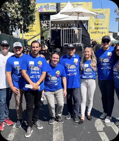 Group of people wearing matching blue event t-shirts standing outdoors in front of a stage with a yellow banner that reads Love Acceptance and Hope.