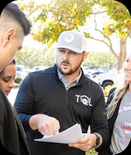 Man wearing a TQL cap and jacket explains paperwork to a small group outdoors under a tree.