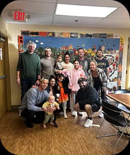 Group of adults and children posing indoors against a wall decorated with photos and colorful items.