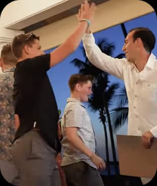 A man in a white shirt high-fiving a young boy while other boys stand nearby against an evening sky with palm trees.